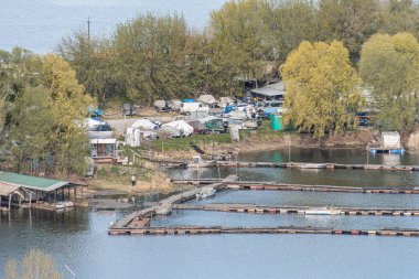 Huzurlu göl manzarasındaki ahşap rıhtım, farklı büyüklükteki teknelerin ışık kümesini yansıtan kayıkhaneler çatıları olan küçük yapılar sakin göl suyu Kyiv, Ukrayna 04-19-2025