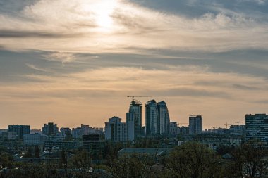 Yapılar, ağaçlar ve uzun gölgeli gölgeli şehirler. Beton yapılar, vinçler ve kentsel gelişime dair ipuçları. Baskın renkler Kyiv, Ukrayna 04-19-2025.