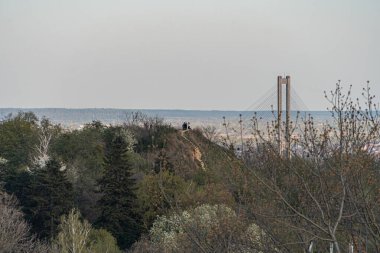 Huzurlu bir manzarada nehir veya vadi üzerinde çok kemerli bir köprü, birbirine bağlı yolda uzak araçlar, kümelenmiş binalar ve çeşitli bitki örtüsü Kyiv, Ukrayna 04-19-2025 yer alıyor.