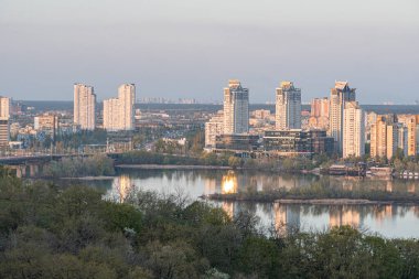 Alacakaranlıkta yüksek binalar, çeşitli mimari stiller, yeşillik, su yansıması, uzak ağaçlar ve karşı taraftaki Kyiv, Ukrayna 04-19-2025 bankasındaki küçük bir bina ile kentsel ufuk çizgisi