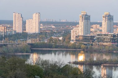 Çok sayıda yüksek konutlu modern şehir manzarası, durgun su yansıtan binalar, köprü, sessiz toprak tonları, yumuşak ışıklandırma ve yeşil bitki örtüsü Kyiv, Ukrayna 04-19-2025