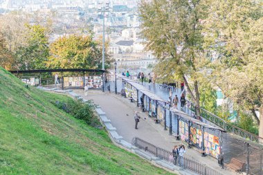 Çimenli verimli tepeli şehir parkı, metal çitlerin üzerindeki renkli kıç işaretleri, karışık mimari ile şehir silueti, bir kentin sanat ve doğasını yakalayan samimi an Kyiv, Ukrayna 10-11-2024