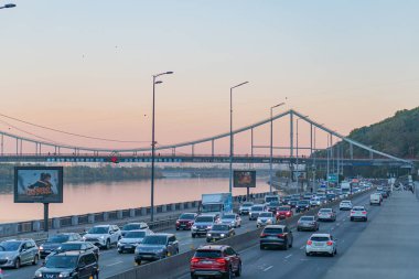 Altı şeritli otoyol, hareket halindeki arabalar, mavi köprü, sakin su, alacakaranlık, kemerli köprü ışıkları, dağ sırası, pembe, turuncu gökyüzü Kyiv, Ukrayna 10-22-2024