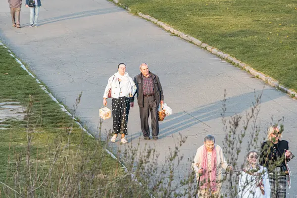 Bir grup insan açık havada kaldırımlı bir yolda yürüyor, parkı kuruyor, çeşitli giysiler giyiyor, bazıları siyah beyaz benekli ceket, koyu renk elbise, eşarp B Kyiv, Ukrayna 04-19-2025