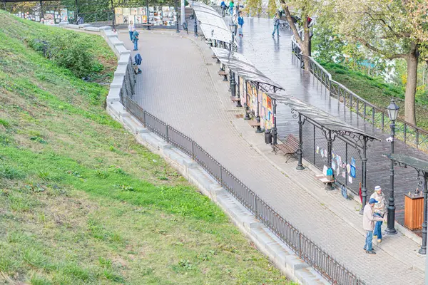 İnsanlar, banklar, ağaçlar, çimenli alanlar, ticari binalar ve arka planda Kyiv, Ukrayna 10-11-2024 tabelasının yer aldığı panoramik bir açık hava sahnesi