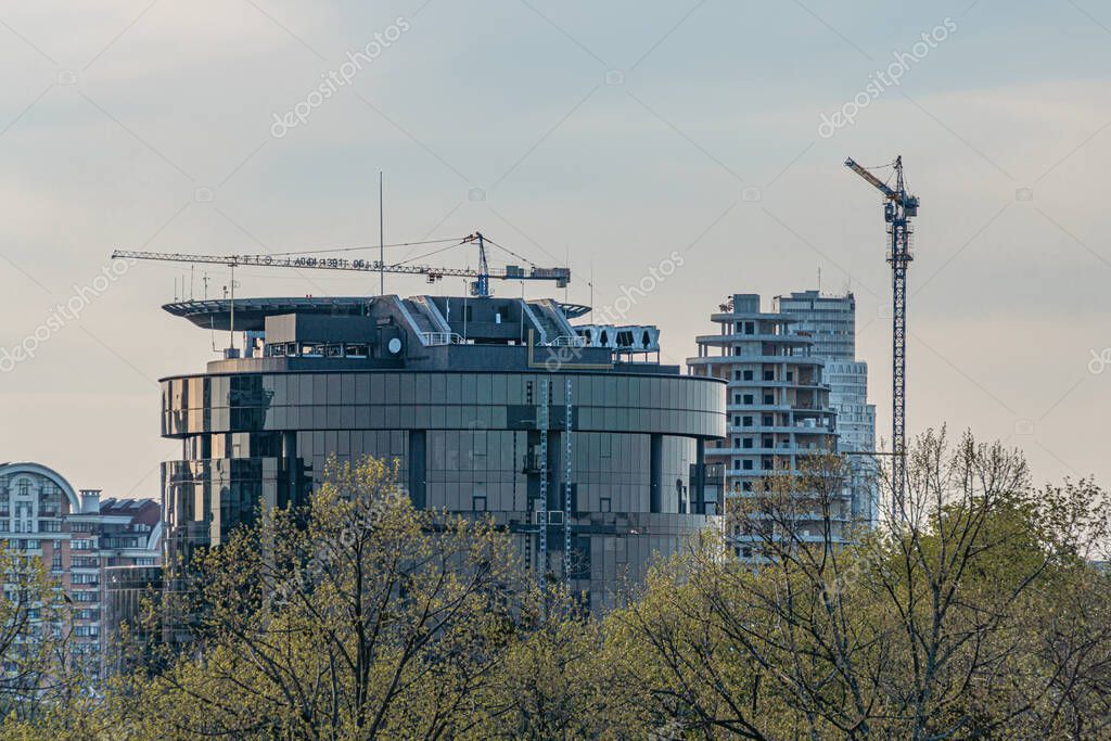 Twilight cityscape with tall, varied buildings, prominent construction cranes, business district setting, deep blues and grays, no people or moving vehicles, waterma Kyiv, Ukraine 04-19-2025