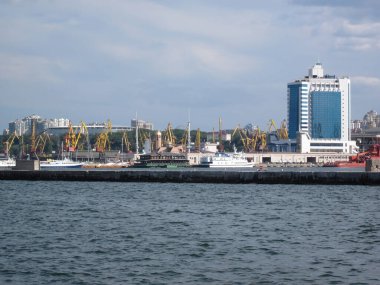 Modern bina, endüstriyel yapılar, vinçler ve küçük beyaz bir teknenin bulunduğu liman sahnesi rüzgarlı bir günü işaret ediyor. Parçalı bulutlu gökyüzü, gerçekçi stil, natura Odessa, Ukrayna 06-07-2012