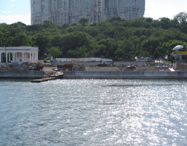 Rıhtım sahnesinde solda inşaat halindeki klasik bir bina, suyun karşısında bir apartman kompleksi, ön planda küçük bir tekne ve pürüzlü ışıklandırma - Odessa, Ukrayna 06-07-2012.
