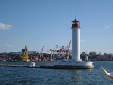 Deniz feneri ve nakliye konteynırları, bulutlu gökyüzü altında, sakin su altında, geleneksel tasarım, kırmızı kubbe, yakınlardaki sanayi bölgesi, sessiz renkler, yumuşak ışık - Odessa, Ukrayna 06-07-2012.