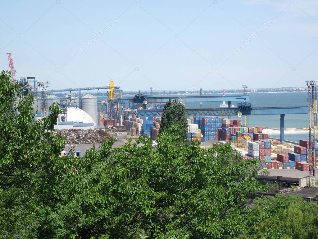 Busy port scene under overcast sky with patches of blue and scattered clouds, lush green trees in foreground, stacked shipping containers white, yellow, blue, orang Odessa, Ukraine 06-15-2012