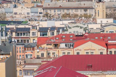Kırmızı ve kahverengi binalar, karışık mimari tarzlar, çatılarda güneş panelleri, arka planda kayan tepeler, bulutlu hava, yüksek perspektiften panoramik manzara