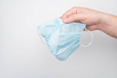 Buruşuk bir açık mavi cerrahi maske, tarafsız bir iç mekan arka planına karşı elle tutulur. Maskenin üç pili ve iki kulağı vardır. Doğal ışıklandırma yumuşak gölgeler bırakır..