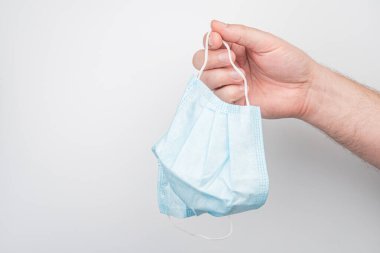 Merkezli bir el, açık mavi cerrahi bir maskeyi arka planda sade bir arka planda tutarken yapay ışıklandırma içeriden bir ayar izlenimi veriyor. Ek nesneler veya metinler görünmüyor. Tarz s