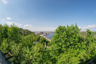Ön planda yemyeşil yemyeşil, sık ağaç ve çalı örtüsü, geniş su gölü veya sakin nehir gövdesi, parçalı bulutlu gökyüzü, uzaktaki şehir manzarası, yumuşak gündüz kuşağı.