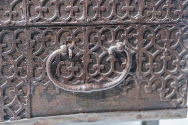 Karmaşık metal işlemeli antik sandık, koyu ahşap, süslü çiçek desenleri, merkezi yuvarlak saplı, hafif yıpranmış metal elementler, nötr arka plan.