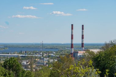 Doğal ve inşa edilmiş ortamların karışımına sahip bir sanayi bölgesinin hava manzarası. Sahne çimenli bir tepe, temiz gökyüzünü yansıtan bir su kütlesi, kırmızı ve beyaz şeritli faktoryum depoları içeriyor.