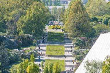 Bulutlu park manzarası asfalt yol, yemyeşil çimenler, ağaçlar, günlük kıyafetler içindeki insanlar, büyük bina, yumuşak ışık muhtemelen yüksek bir yerden alınmış.