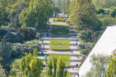 Uçsuz bucaksız şehir parkı asfalt yol, yemyeşil çimen, olgun ağaçlar, modern bina, banklar, insanlar, bulutlu gün, yumuşak ışık, kuş bakışı görüş, görünür metin yok