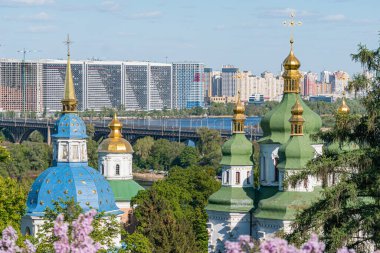 Mavi ve yeşil çatılı iki ayırt edici Doğu Ortodoks binası, altın soğan kubbesi, ön planda yemyeşil, arka planda modern mimarisi olan şehir manzarası, açık gökyüzü.