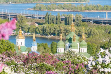 Ön planda birden fazla kubbesi olan kilise kompleksi bulunuyor, en büyüğü süslü ve altın aksanlı, çiçek açan ağaçlarla çevrili köprü, arka planda su yansıtan sahne boyunca durgun.