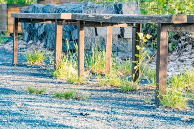 Açılı ahşap bank, çakıl zemini, seyrek çimen yamaları, kayalık duvar arkaplanı, sıcak ışıklandırma, öğleden sonra geç saatlerde