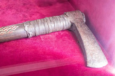 Kapağında karmaşık desenler olan, aşınmış metal bir çekicin yakın plan görüntüsü, kırmızı kadife kaplı bir çantada, kapalı bir müzenin arka planında, yumuşak iç mekan aydınlatması altında sergileniyor.