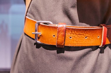 Gümüş metal çivili ve dikdörtgen tokalı kahverengi deri kemerin yakın plan görüntüsü, koyu renk giyinen biri tarafından giyilen kahverengi-beyaz bir arka plan.