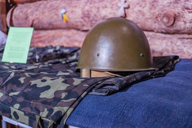 Askeri teçhizat sergileniyor. Koyu yeşil dış görünüş, zeytin rengi, kamuflaj kumaşı düzensiz şekiller, ağaç yaprağı gibi dizayn, koyu yeşil ve bronz renkler, açık hava ayarı kayıtları veya ahşap panel