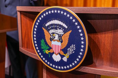 Resim, Beyaz Saray 'da mavi bir kürsüye odaklanmış bir basın toplantısını tasvir ediyor. Bir kartal amblemi, oklar, zeytin dalı, yıldızlar ve çizgiler mevcut.