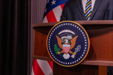 Amerikan bayrağı ve başkanlık mührü sergilenen bir hükümet ayarı. Resmi giyinmiş bir şahıs kürsünün arkasında duruyor. Muhtemelen konuşmaya hazırlanıyor ya da Natur 'u yeni bitirdi.