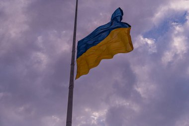 Bulutlu gökyüzünde Ukrayna bayrağı dalgalanmakta, yüksek bir yapıya bağlı, görülebilir bir zemin ya da insan figürü yok aydınlatmalar öğleden sonra erken saatlerde düz fotografı gösteriyor.