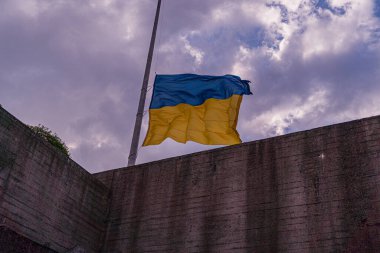 Bulutlu gökyüzünde Ukrayna bayrağı dalgalanmakta, yüksek bir yapıya bağlı, görülebilir bir zemin ya da insan figürü yok aydınlatmalar öğleden sonra erken saatlerde düz fotografı gösteriyor.