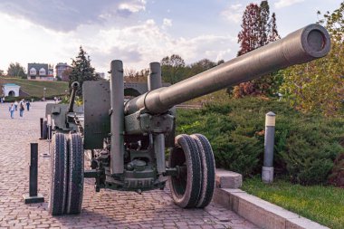 Park ortamında büyük bir askeri top, tekerleklerde siyah bir fıçı ve alevli bir ağızlık, dağılmış ağaçlar, kaldırım taşı zemin, açık alan, parçalı bulutlu gökyüzü, uzak mesafede birkaç kişi.