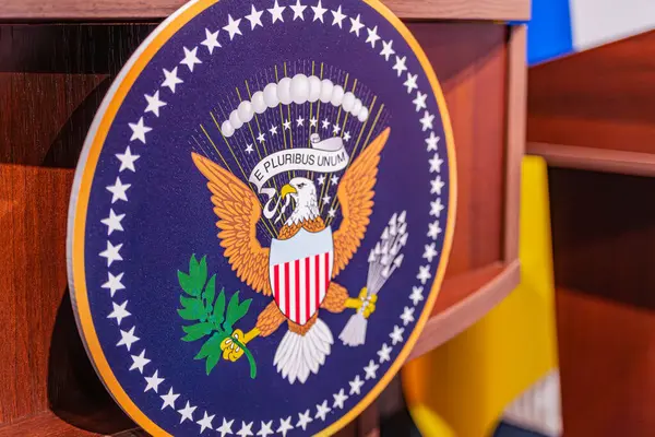 Bir ayağı zeytin dalında, diğer ayağı da bir kartalın üzerinde olan yuvarlak bir ABD ambleminin yakın plan fotoğrafı. Amblem, kartalların kafasının üzerinde ve Birleşmiş Devletler 'in üzerinde bir dairede 50 yıldıza sahiptir.