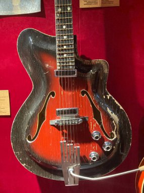 Kırmızı kadife bir yüzey üzerinde f-deliği olan kırmızı elektro gitar, karanlık bir arkaplan karşısında sergilenen ek nesneler ve muhtemelen ilgili bilgiler içerir Kyiv, Ukrayna 06-19-2022