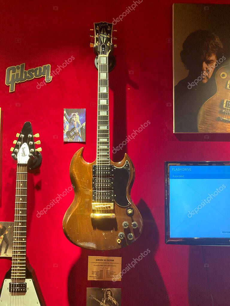 Focus on two guitars against a dark backdrop, one with a bands logo pickguard, in an exhibit or museum setting, showcasing additional guitars slightly out of focus Kyiv, Ukraine 06-19-2022