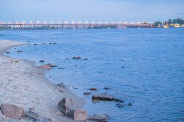 Alacakaranlık sahnesi sakin bir nehir portakalı mavi gökyüzüne yansıtan, altın dalgalar, kayalık kıyı şeridi, su altındaki kayalar, köprü yapıları, doğal ortam.