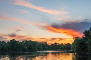 Günbatımı göl manzarası turuncu ve pembe gökyüzü, sakin göle yansıyor. Önplanda siluetli ağaç, insan ya da yapı yok..
