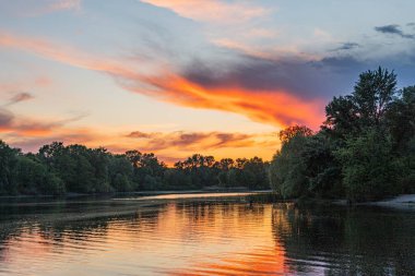 Geniş bir nehir üzerinde gün batımı sahnesi. Portakal, pembe ve koyu gökyüzü. Dağılmış bulutlar. Sakin su yüzeyi günbatımı renklerini yansıtıyor.