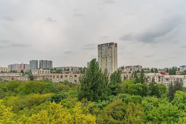 Sağında uzun, modern bir bina ve solunda canlı ağaçlar olan sakin, bulutlu bir şehir manzarası. Görüntü, kentsel mimarinin uyumlu bir harmanını sunuyor.