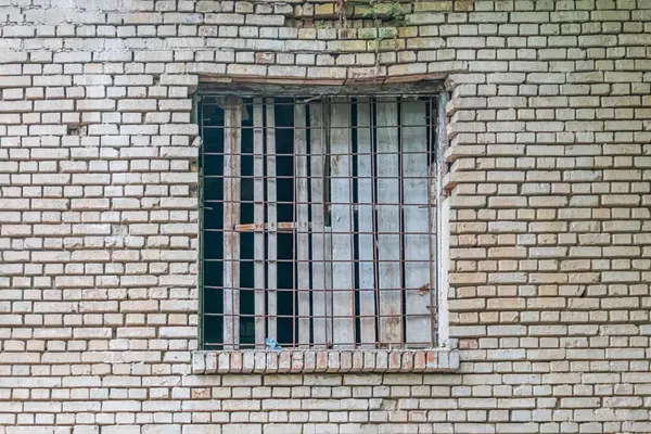 Eski bir tuğla binanın yakın görüntüsü, metal parmaklıklı pencere, yıpranmış tuğlalar, yumuşak gölgeler, bastırılmış atmosfer, sessiz renkler, doğrudan bakış açısı.