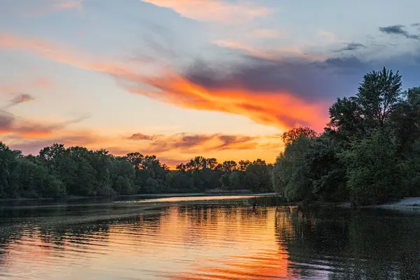 Geniş bir nehir üzerinde gün batımı sahnesi. Portakal, pembe ve koyu gökyüzü. Dağılmış bulutlar. Sakin su yüzeyi günbatımı renklerini yansıtıyor.