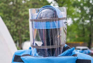 Gaz geçirmez, koruyucu kask ve takım elbise giyen biri gün ışığında sokakta dikiliyor. Arka planda ağaçlar ve park edilmiş araçlar var.