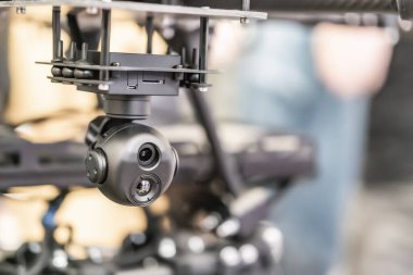 Endüstriyel çalışma alanı içinde çalışan bir robotik kola bağlı bir savaş kuadkopter dronu gösterilir. Kol, uç kapağında küresel bir kamera montajı ile orta soldan yukarı doğru hareket eder.
