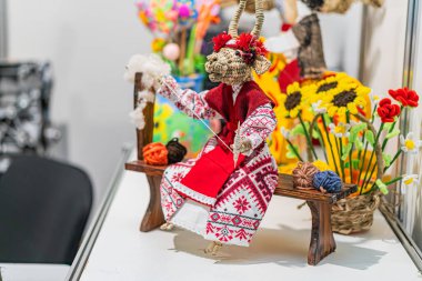 Canlı bir sahnede, kırmızı ve beyaz desenli özenle hazırlanmış bir kuş figürü sergileniyor. Çiçek dolu bir sepet ile ahşap bir ekranda duruyor.