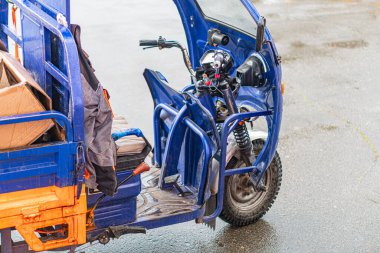Elektrikli kargo üç tekerlekli bisiklet ve motosiklet, bulutlu gökyüzünün altında park edilmiş mavi kurye minibüsü, açık bölmeli nesneleri gösteriyor. Islak zemin, yağmurun yakın zamanda geldiğini gösteriyor.