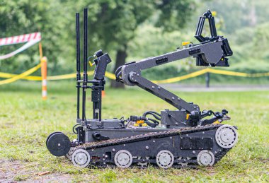 Arka planda ağaçlar olan çimenli bir alanda çok tekerlekli, siyah ve gümüş renkli bir robotik cihaz tasvir edilir. Ağır makinelere benzeyen robotun sensörleri ya da kameraları var.