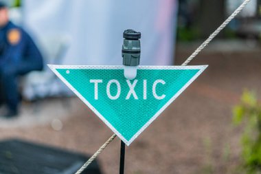 Siyah tel çitin üzerindeki elmas şekilli TOXIC işaretinin yakın plan resmi, çitin arkasında belirsiz figürler olan doğal yeşil bir ortamda.