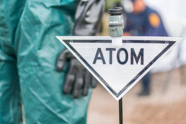 Koruyucu giysili bir kişi açık çimlerin üzerinde bir ATOM tabelasının yanında duruyor. Şahıs açık yeşil bir giysi, kapüşon ve eldiven giyiyor, muhtemelen tehlikeli materyal operasına katılıyor.