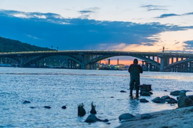 Kaya kıyısında alacakaranlıkta balık tutan kişi, Kiev Köprüsü 'ndeki Dnepr Nehri' nde nehrin üzerinde birden fazla kavis ve sıcak ışıklarla turuncu ve pembe gün batımı gökyüzü yansımaları su yüzeyinde gerçekçi bir şekilde yansıyor.